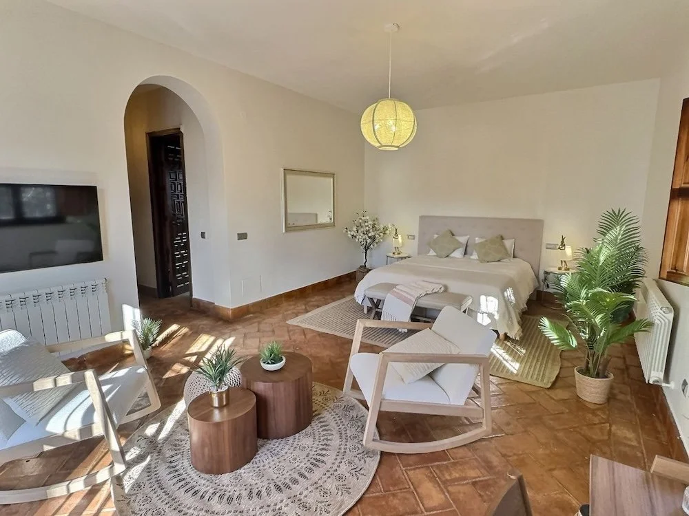 Aitana retreat - Living room with beige walls, tiled floor, and a king-sized bed with beige headboard, pillows, and a blanket. There is a small white armchair, a rocking chair, potted plants, a mirror, and a hanging paper lantern light fixture.