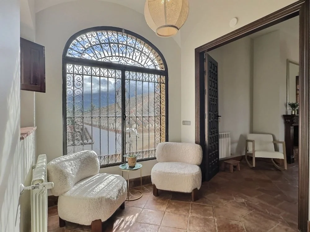 Aitana retreat - Sunlit room with two cream-colored, plush upholstered chairs, a small glass-top side table with a potted plant, a large arched window with ornate black wrought iron bars, a pendant ceiling light.