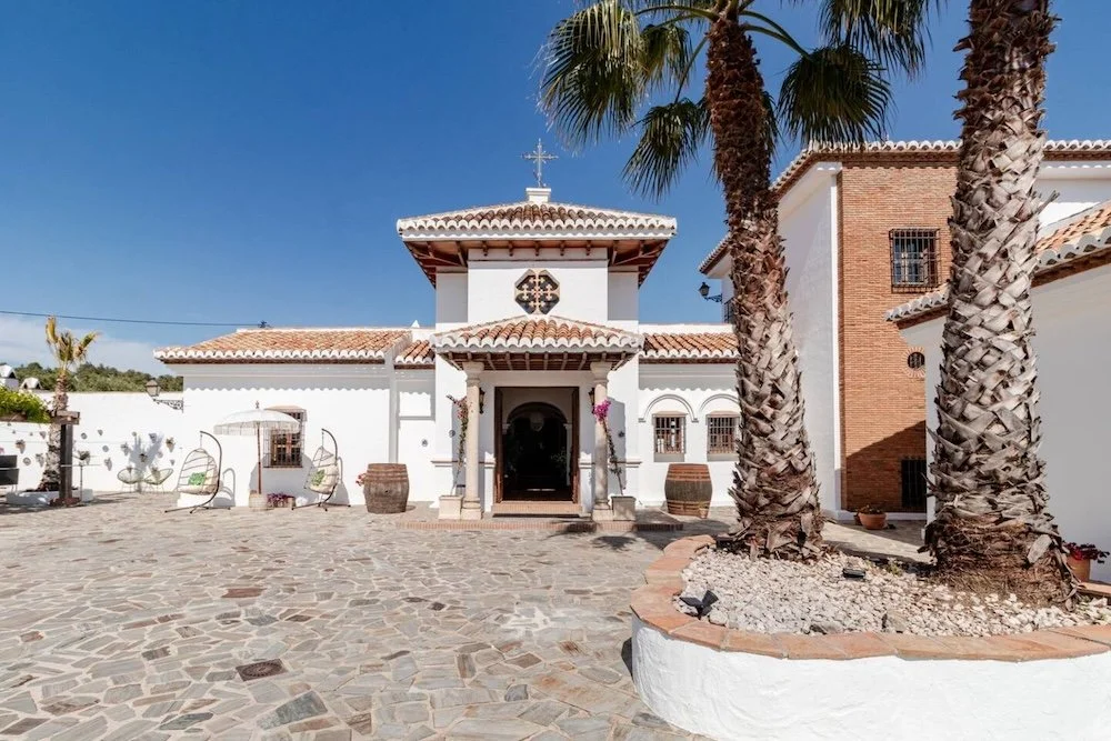 Aitana retreat - A white Spanish-style church with a tiled roof, set in a courtyard with cobblestone paving, two tall palm trees in planter beds, and a clear blue sky.
