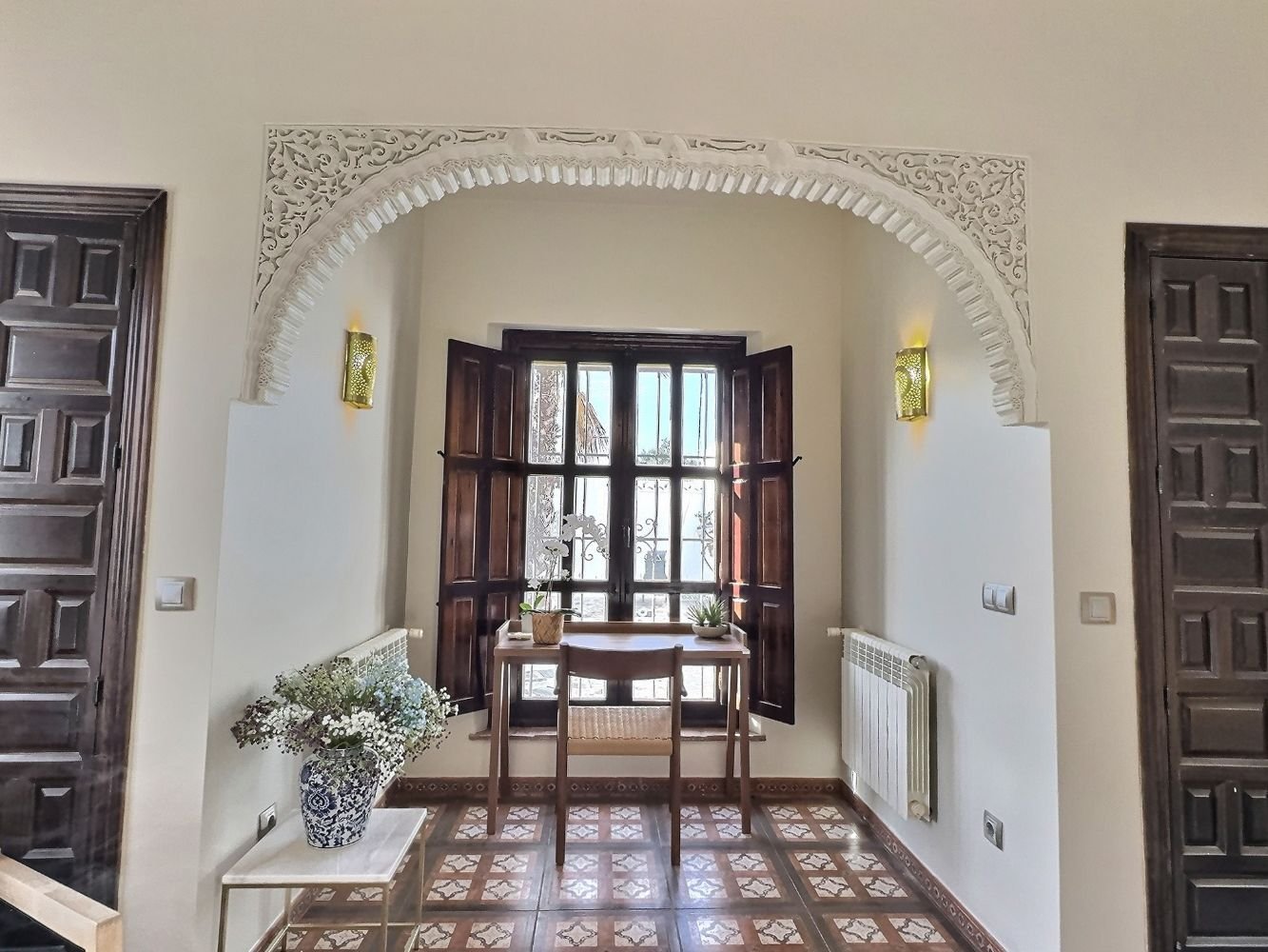 Aitana retreat - Interior view showing a window with open wooden shutters, a small table with potted plants, a chair, a vase with flowers, and decorative lights on the wall.