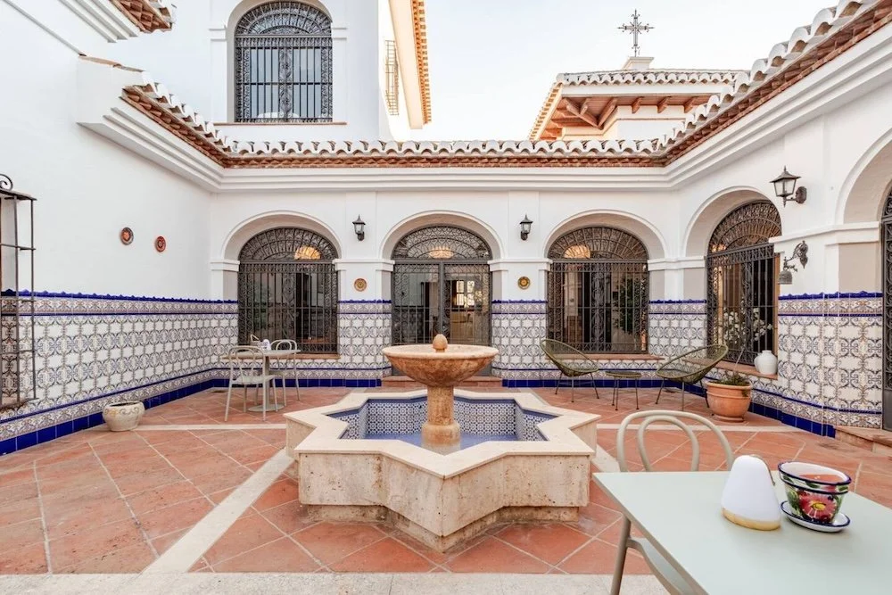 Aitana retreat- Courtyard with decorative fountain, terracotta tiled flooring, and Spanish-style white walls with blue patterned tile accents. There are elegant chairs, potted plants, and barred arched windows.