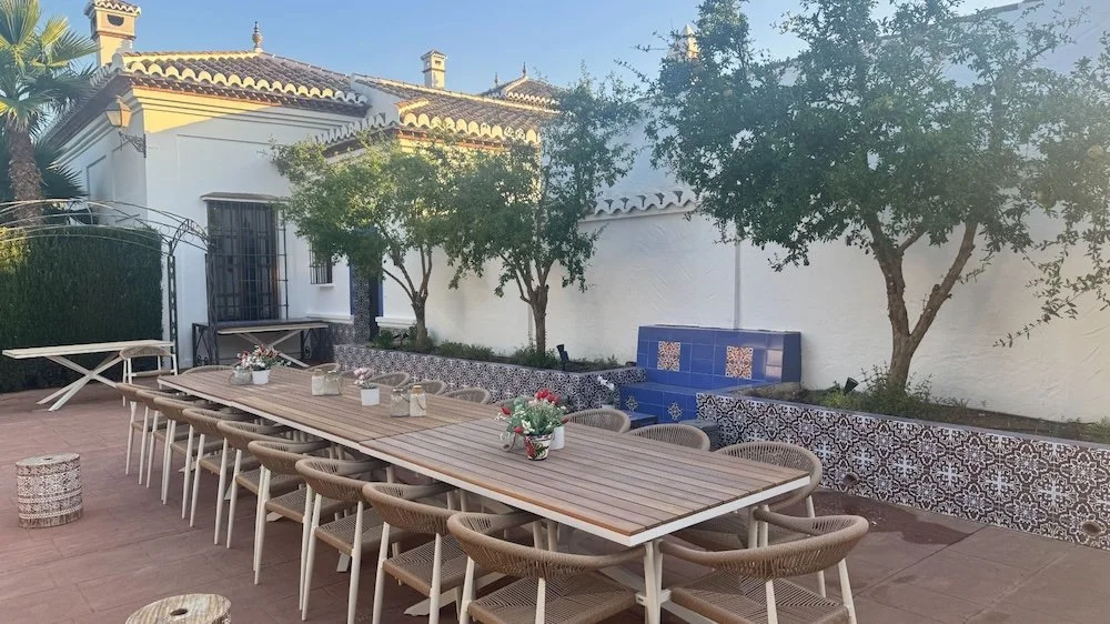 Aitana retreat - Large outdoor dining table with 14 chairs, set on a tiled patio with decorative tiles along the wall, surrounded by trees and a white wall with a yellow and brown house in the background.