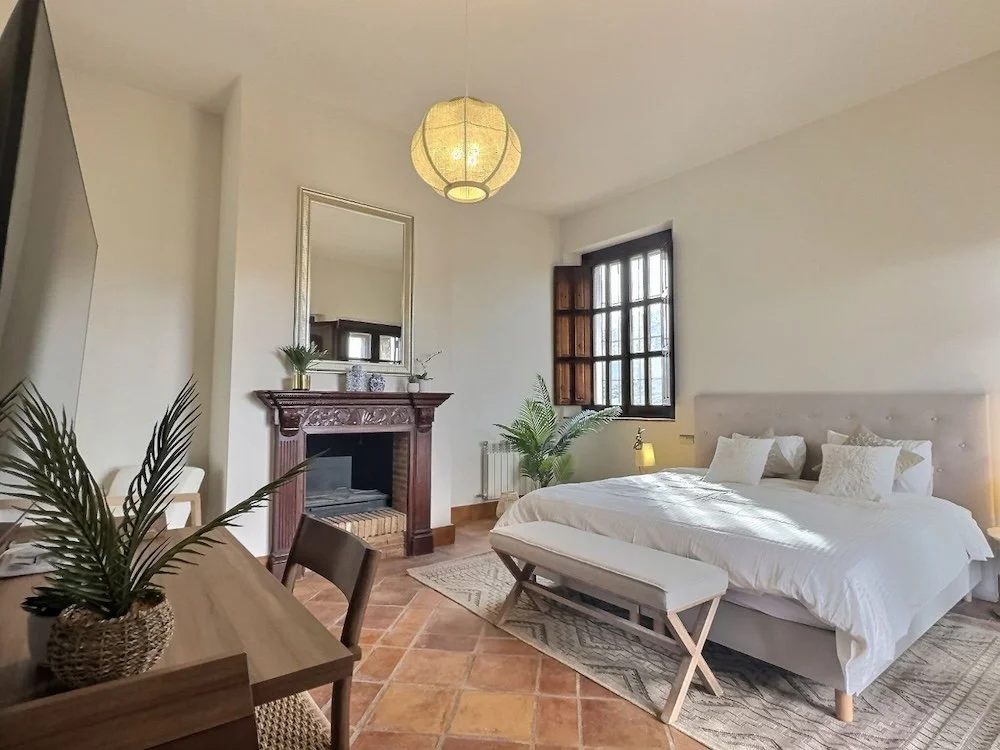 Aitana retreat- A bedroom with a white bed, wooden window shutters, a fireplace with a mirror above it, potted plants, a bedside lamp, and a wooden table with a plant, in a cozy and bright space.