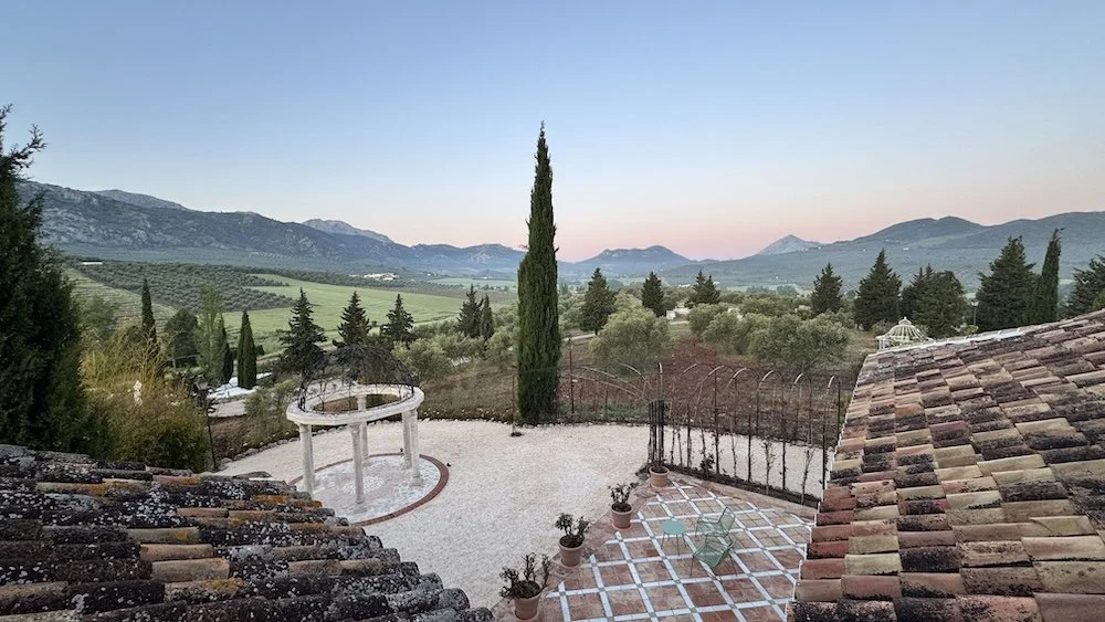 A scenic view of a countryside with rolling hills, scattered trees, and distant mountains during sunset. There is a tiled patio with potted plants and a glass chair, and a white circular structure with columns in the foreground. The scene is viewed f
