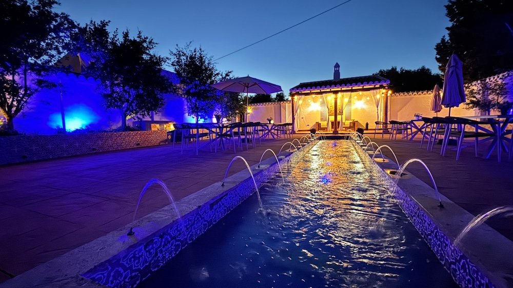 Outdoor patio at dusk with illuminated fountain and seating area, decorated with string lights, umbrellas, and trees.