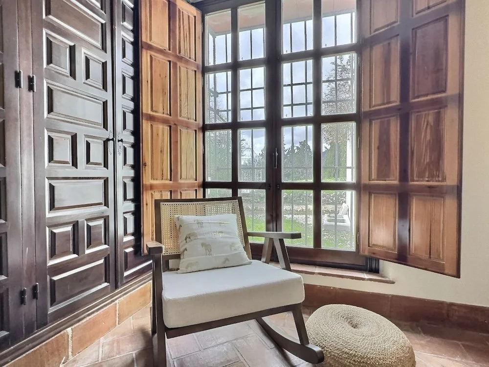 A room with a large window and wooden shutters, a cushioned chair with bamboo backrest, a throw pillow, and a woven ottoman on a tiled floor.