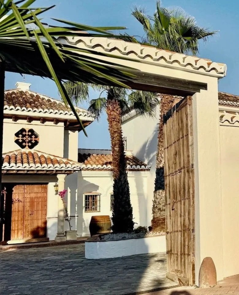 AITANA RETREAT - View of a Mediterranean-style courtyard with a large wooden gate, white stucco walls, red-tiled roofs, and tall palm trees under a clear blue sky.