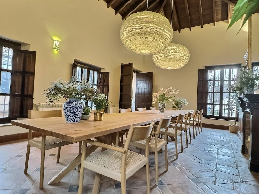 Aitana retreat- A spacious dining room with a long wooden table, eight chairs, floral vases, and large hanging paper lantern lights.