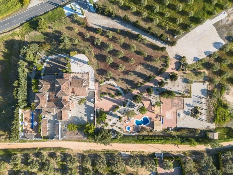 Aitana retreat- Aerial view of a large estate featuring a main house, swimming pool, hot tub, tennis court, and extensive yard surrounded by trees and a dirt road.