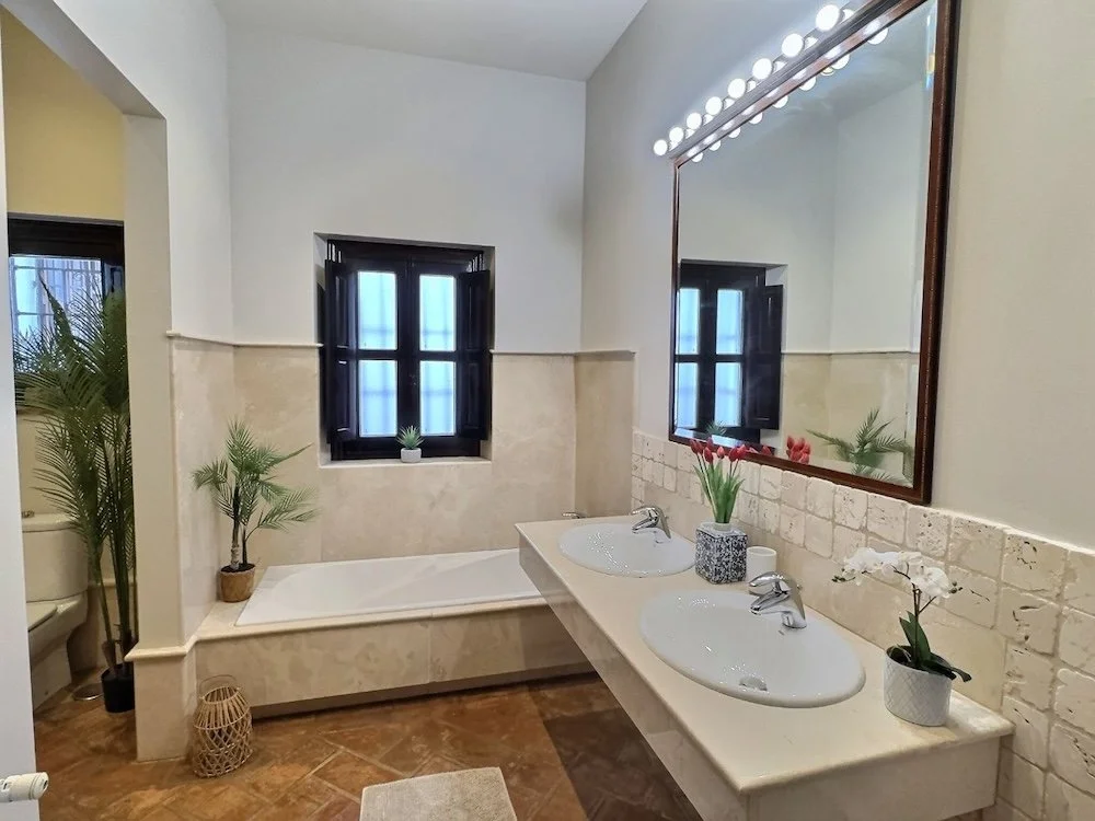 Bathroom with double sinks, large mirror with lights, potted plants, a window, and a bathtub.