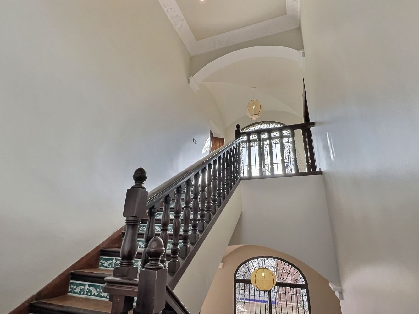 Aitana retreat-Interior view of a multi-story house staircase with wooden handrail, decorative balusters, tiled steps, and arched windows with ornate metal grilles, hanging light fixtures, and white walls.