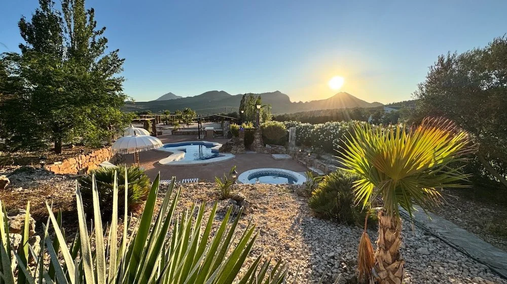 Sunset over a desert landscape with a hot tub, jacuzzi, and lounge area surrounded by desert plants and mountains in the background.