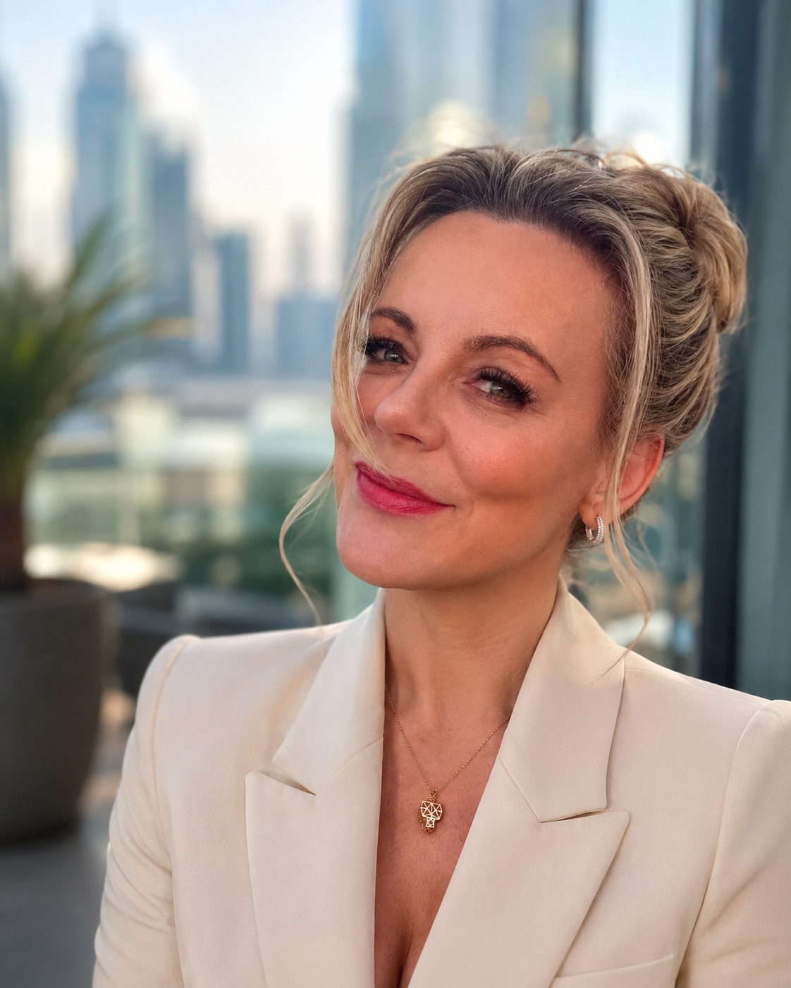 A woman with blonde hair styled in a loose updo, wearing a cream blazer and jewelry, smiling in a high-rise office with city buildings in the background.