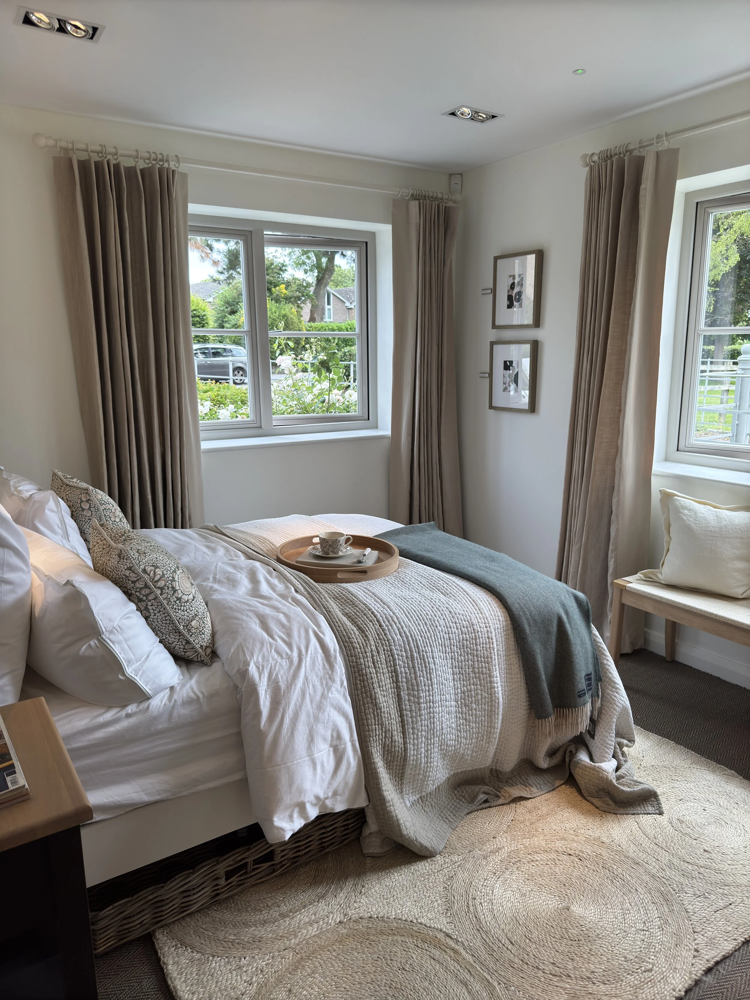 A cozy bedroom with a made bed, beige and white bedding, pillows, a tray with a teacup on the bed, beige curtains, and two framed pictures on the wall, with a window showing greenery outside.