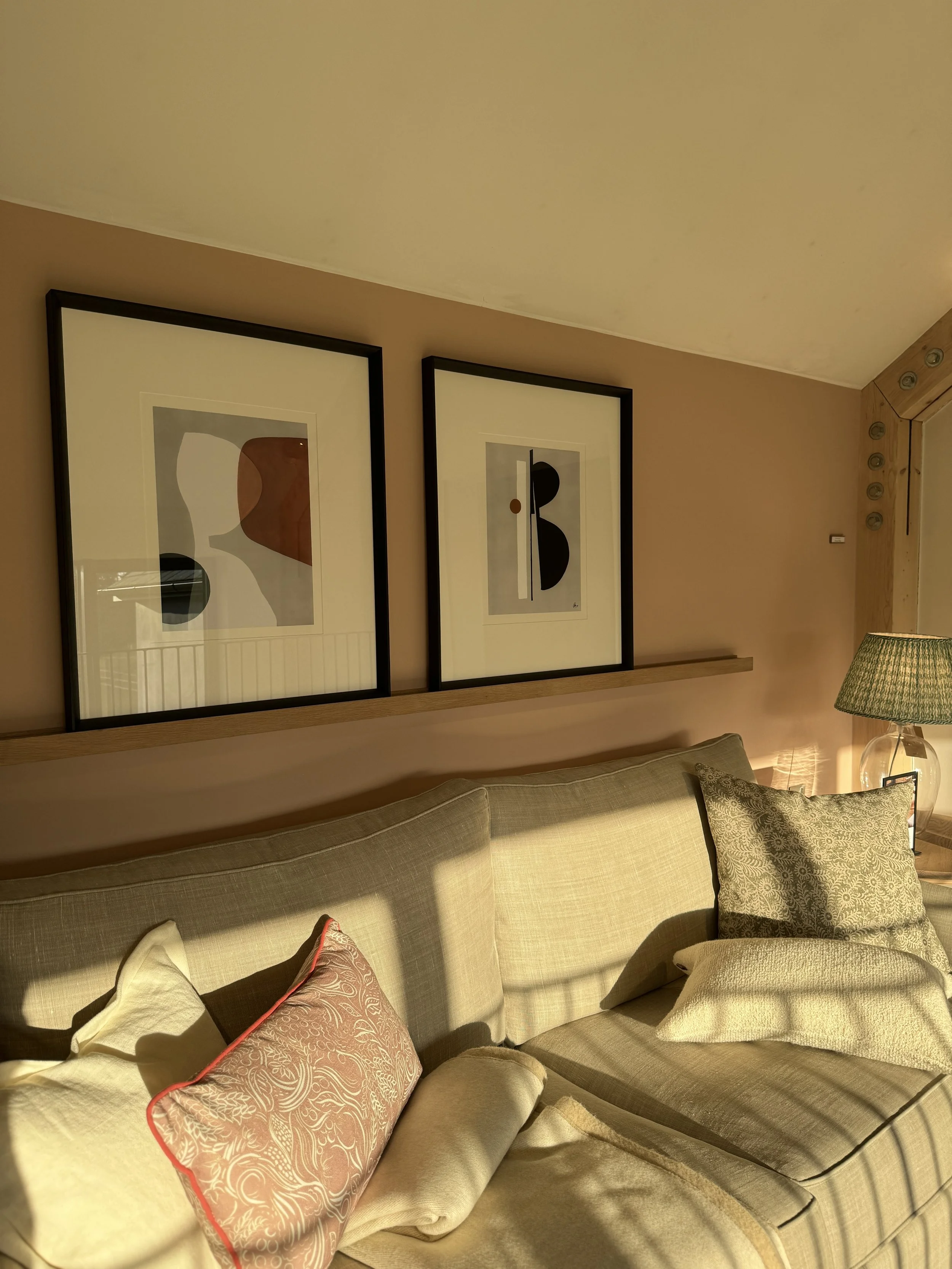 A beige sofa with patterned and plain cushions in front of a wall with three framed abstract art prints, a wooden shelf, and a lamp with a green shade.