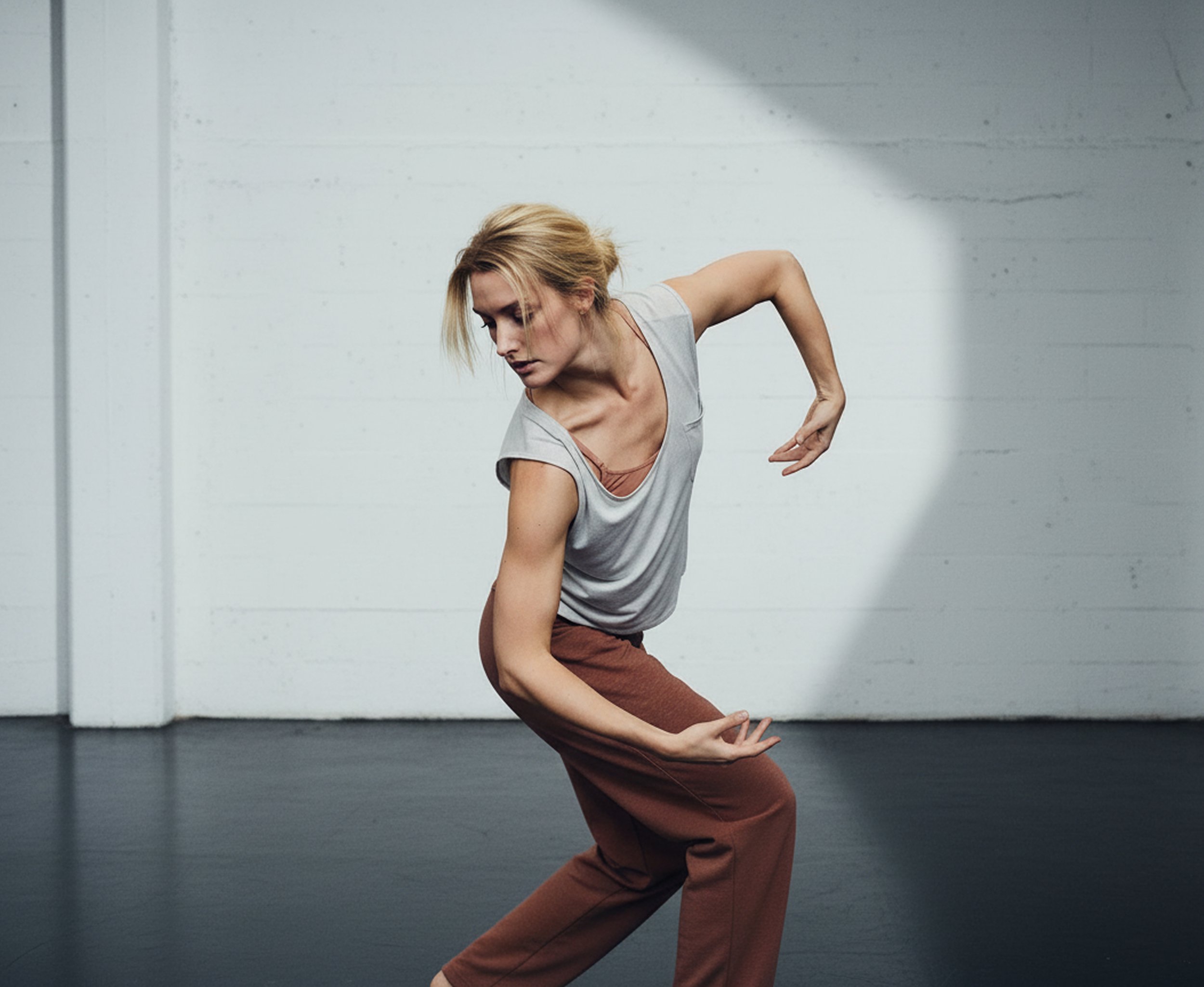 Eine Frau in einer tänzerischen Pose in einem Raum mit weißer Wand und dunklem Boden.