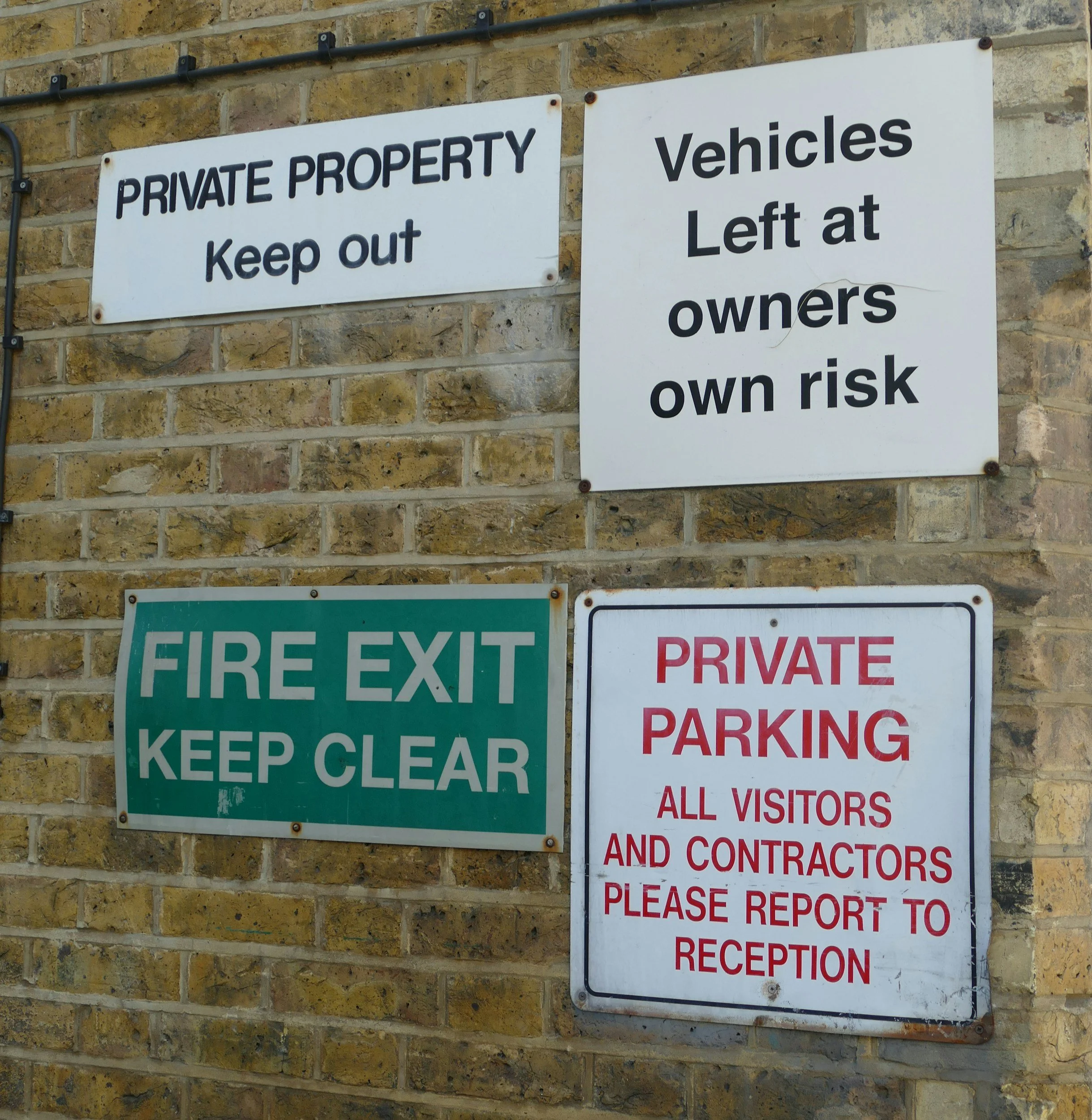 Multiple signs on a brick wall including a 'Private Property Keep Out' sign, a 'Vehicles Left at Owners Own Risk' sign, a 'Fire Exit Keep Clear' sign, and a 'Private Parking' sign instructing visitors and contractors to report to reception.