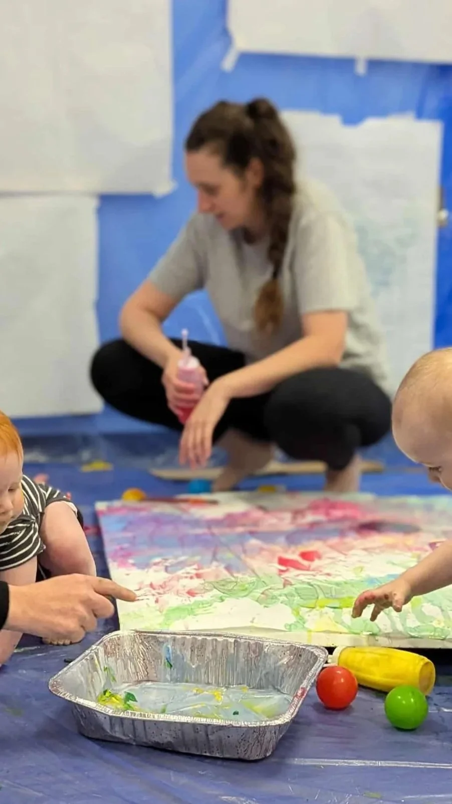 A woman with long brown hair in a braid sitting on the floor with children engaged in finger painting. The woman is holding a squeeze bottle, and children are reaching for paint. Painting supplies and colorful paintings are on the table.