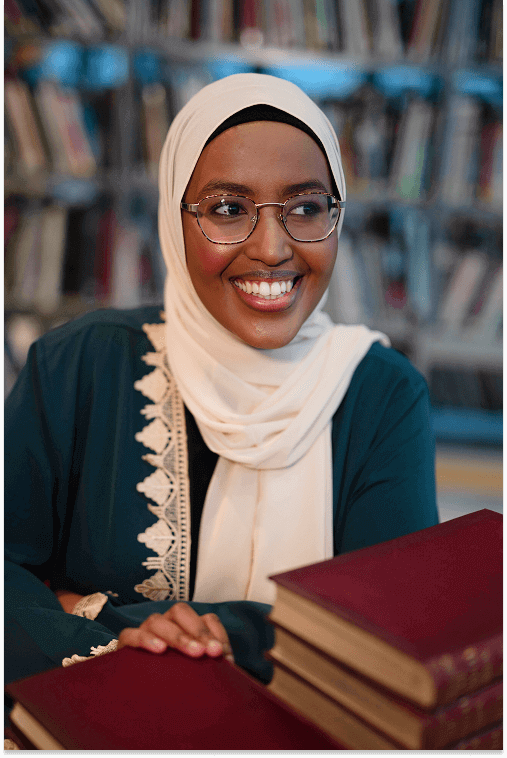 Smiling woman wearing glasses and a white hijab, sitting at a table with a stack of books in a library.
