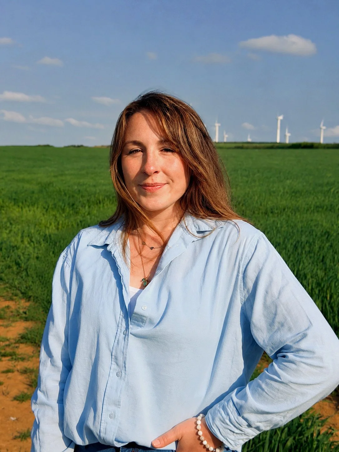 Woman with brown hair wearing a light blue button-up shirt standing outdoors in a green field with wind turbines in the background under a partly cloudy sky.