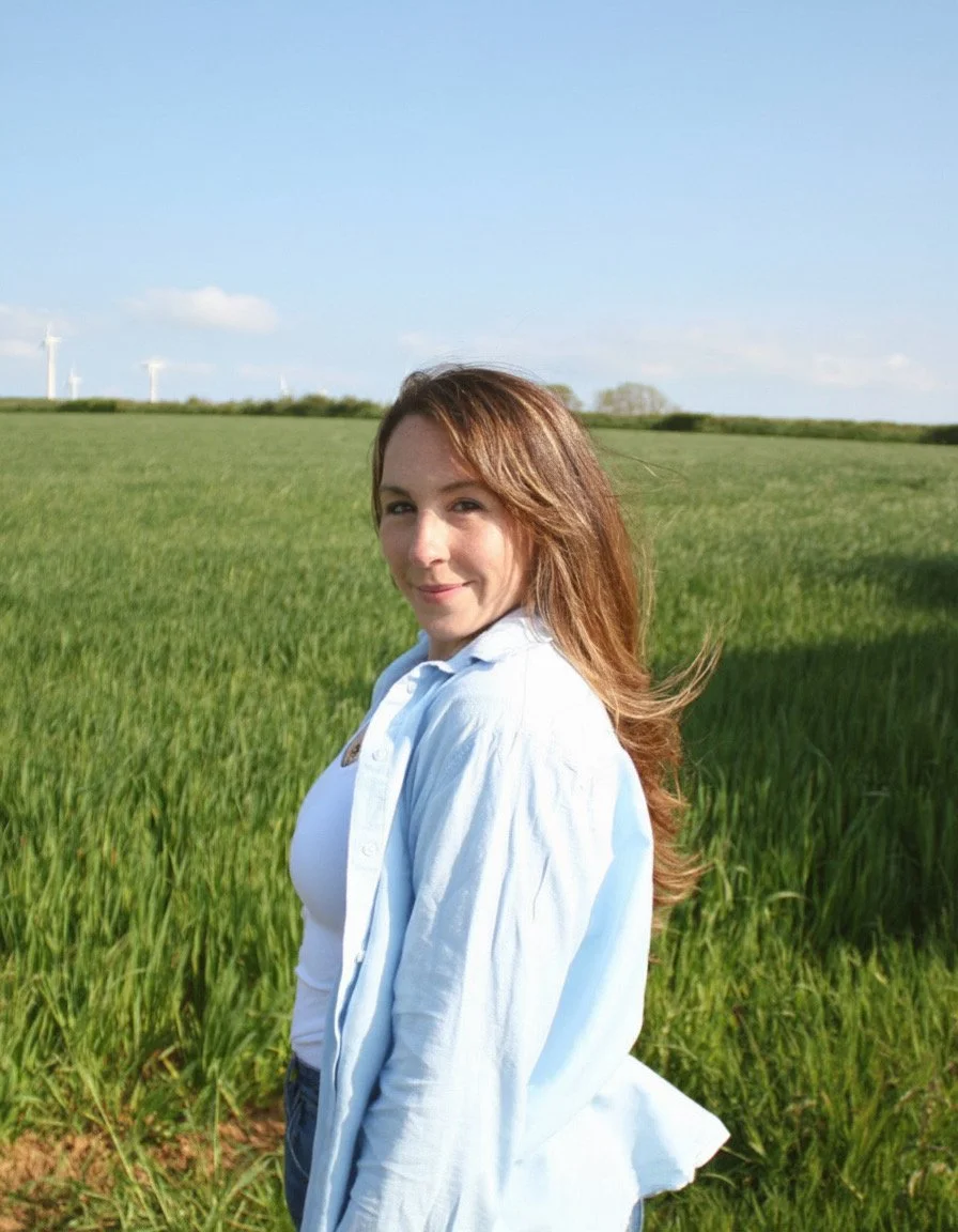 A young woman with long brunette hair wearing a white top and light blue button-up shirt standing in a green field on a sunny day with wind turbines in the background.
