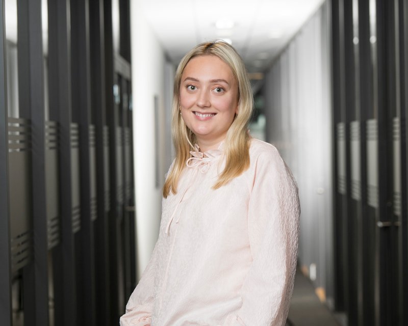 En ung kvinne med blondt hår smiler i et kontorlandskap.