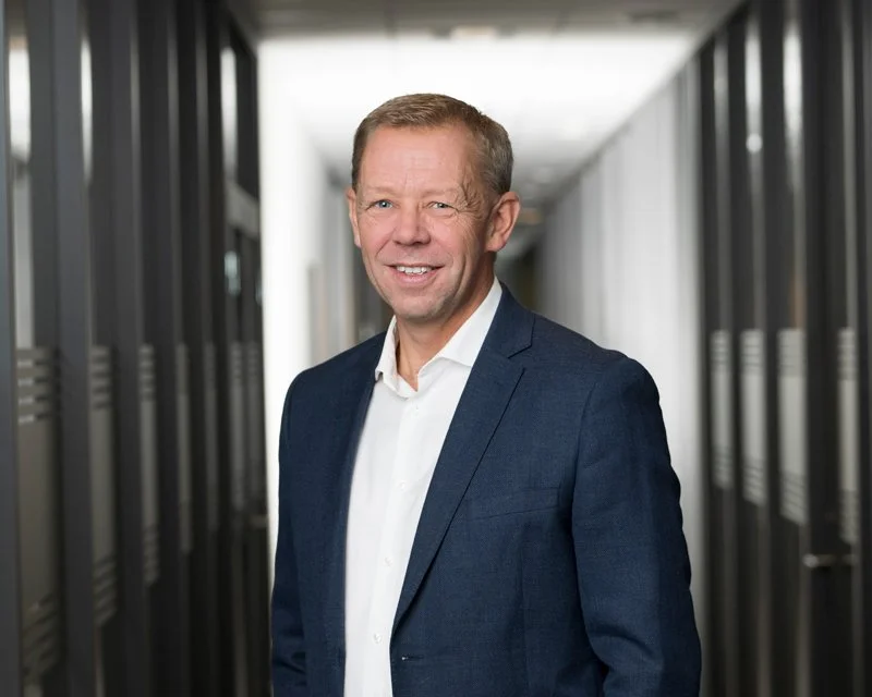 En mann i blå dress og hvit skjorte, smilende, i et kontormiljø med skap på gangen.