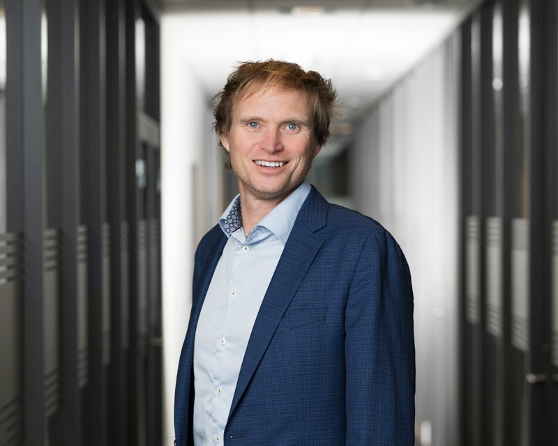 En mann i blå dress smiler og står i en moderne kontorbygning.