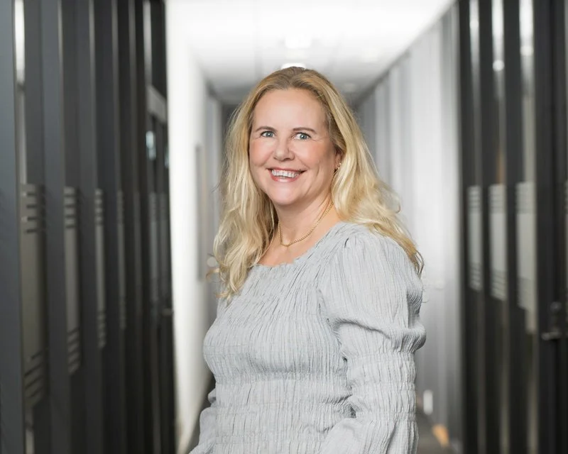 En kvinne med blondt hår smiler i en gang mellom servere i et datarom.