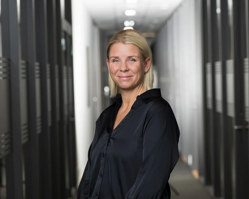 En kvinne med blondt hår iført en svart skjorte står i en korridor med serverskap.