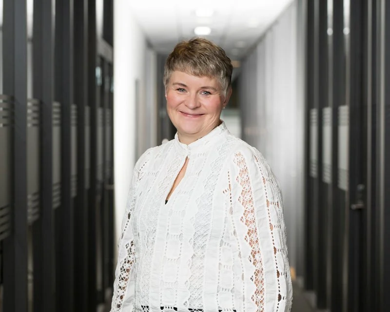 Kvinne med kort blond hår smiler, står i en korridor med serverskap på begge sider.