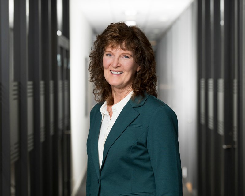 En kvinne med brunt, krøllete hår i en kontorbygning, iført blå dress og hvit skjorte, står foran en rekke serverskåp.