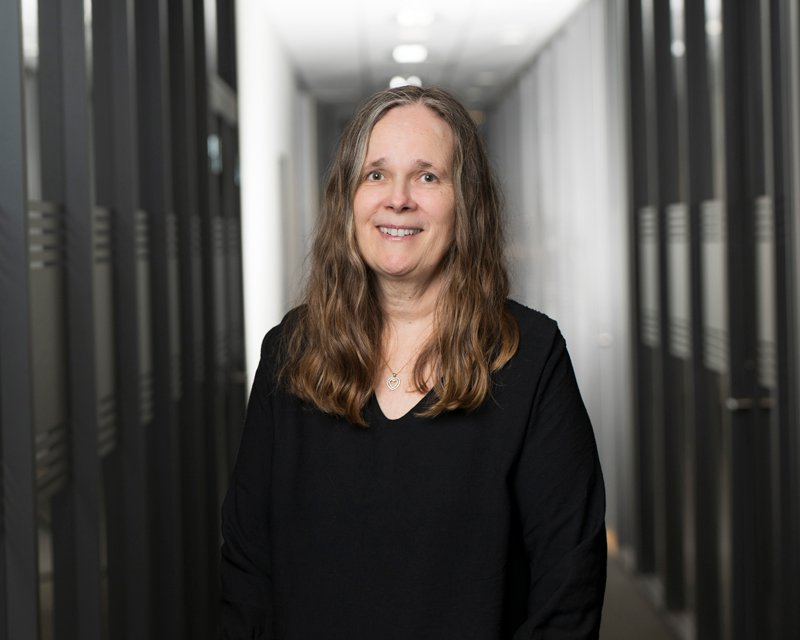 En kvinne med langt, brunt hår som smiler, iført svart t-skjorte, står i en korridor med skap langs væggene.