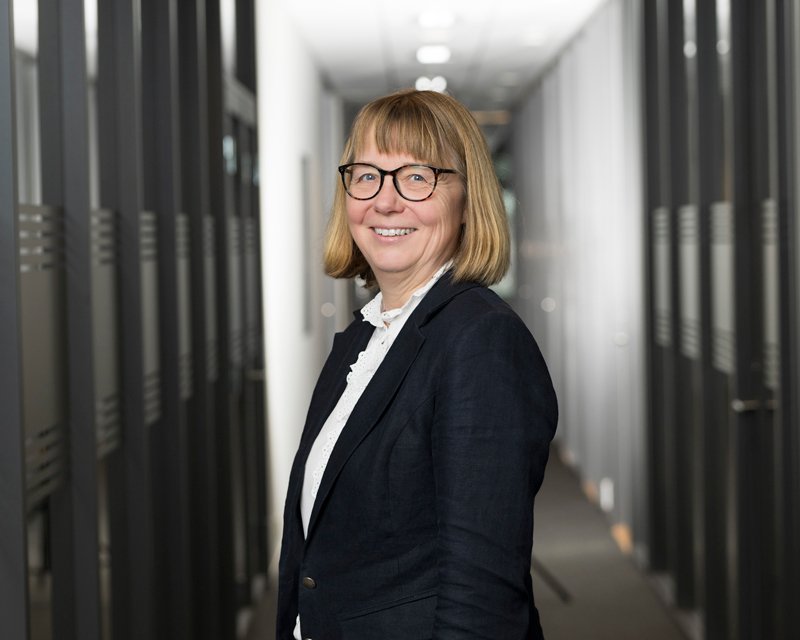 Smilende kvinne med briller, kort blond hår, iført svart jakke og hvit bluse, standing i et kontorbygning med dataservere i bakgrunnen.