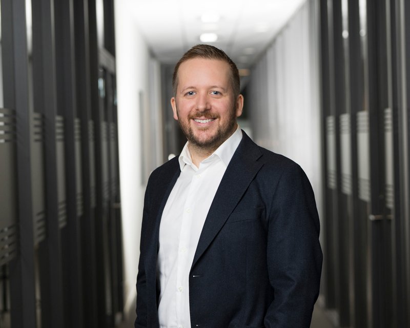 En mann i dress smiler i en kontorbygning.