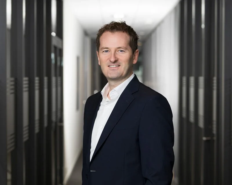 Mann i mørkeblå dress og hvit skjorte står i en gang mellom dataservere og smiler mot kamera.