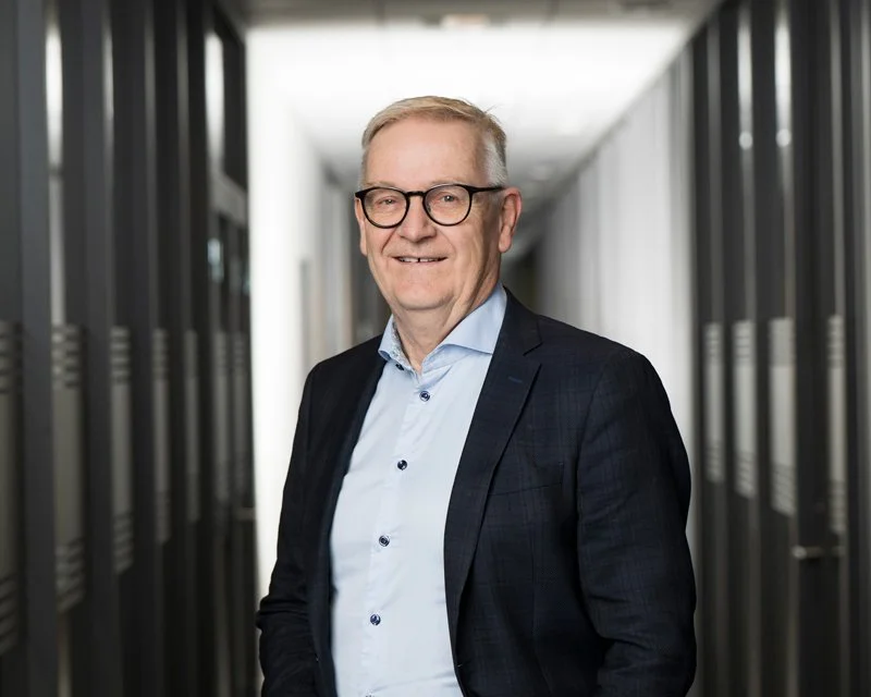 En eldre mann med briller Smiler i et moderne kontorbygg, iført en blå skjorte og blazer.