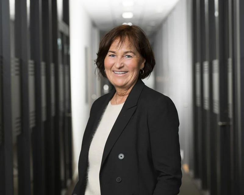 En smilende kvinne med brunt hår i en svart blazer står i et datasenter med serverskåp på begge sider.