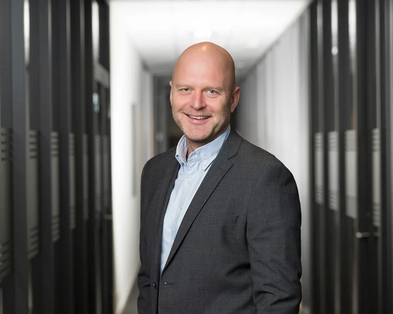 En mann i dress står i en gang mellom dataservere, smilende mot kameraet.