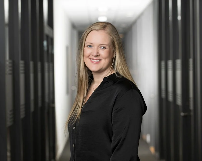 En smilende kvinnelig person med lyst hår ser mot kamera, iført svart skjorte, i en korridor med serverskap på begge sider.