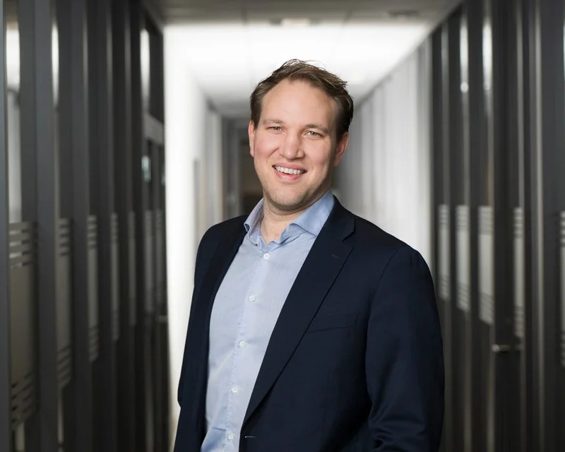 En mann i forretningsantrekk står i en gang mellom dataservere, smiler mot kamera.