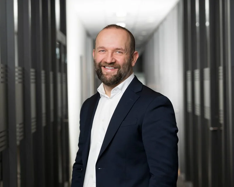 En mann i dress smiler i en kontorbygning.