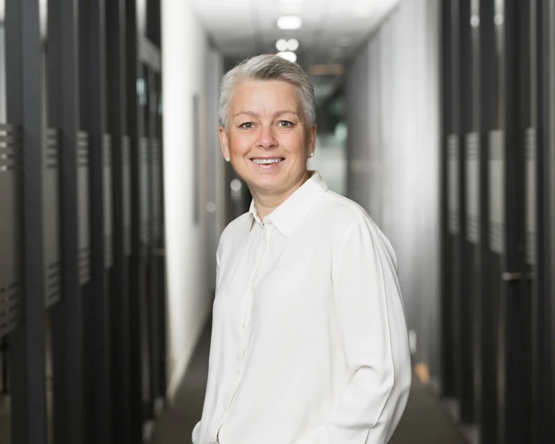 En eldre kvinne med kort, grått hår smiler mens hun står i en serverromgang med serverrack på begge sider.