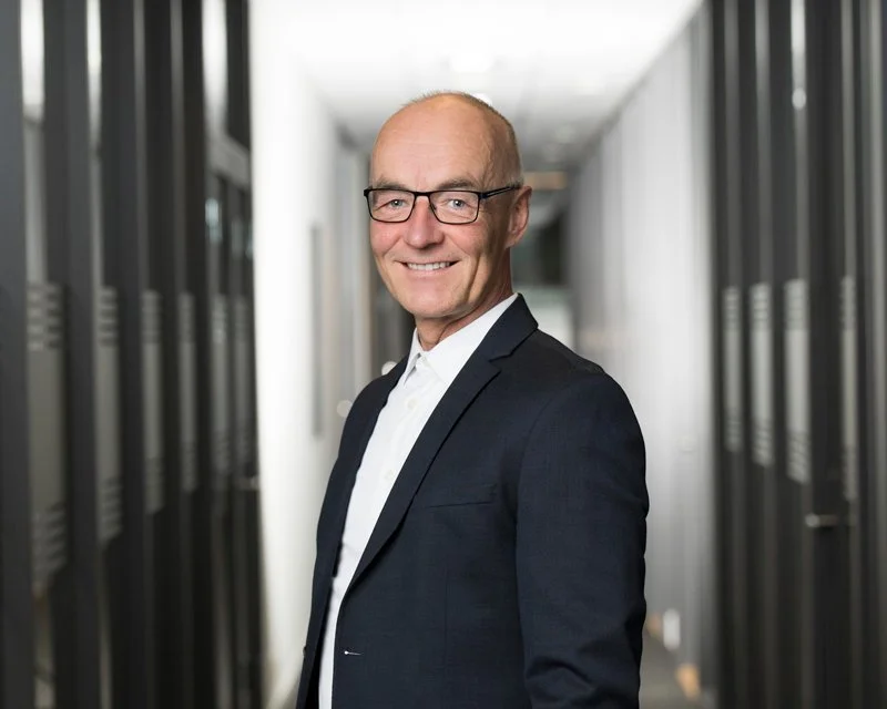 En mann i dress står i en korridor med servere på begge sider, smilende og ser mot kameraet.