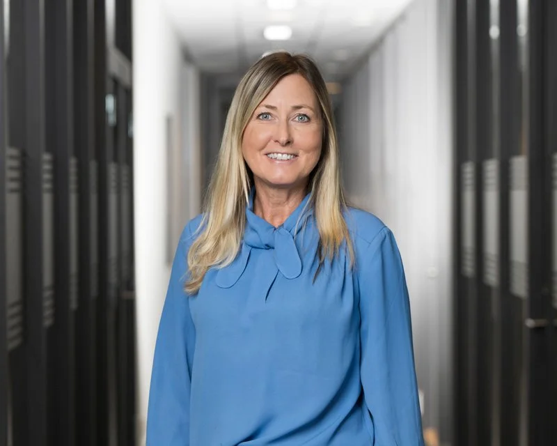 En kvinne med blondt hår og blå bluse står i en datasal med servere.