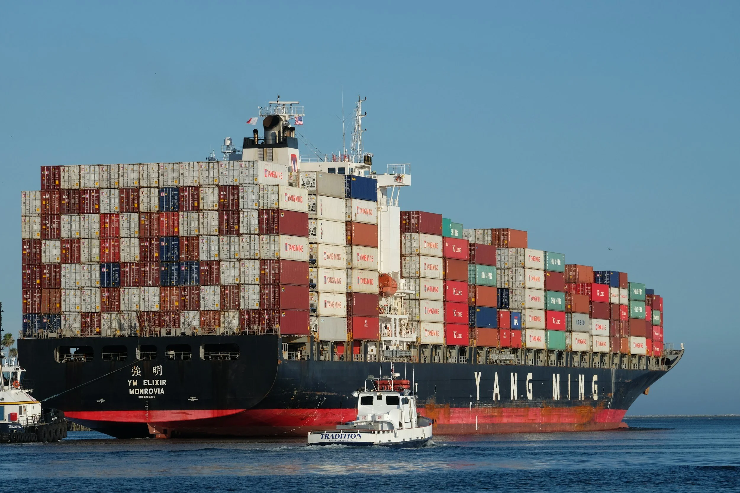Large cargo ship named YM ELIXIR from Monrovia, loaded with numerous colorful shipping containers, sailing in a calm body of water with a smaller boat nearby.