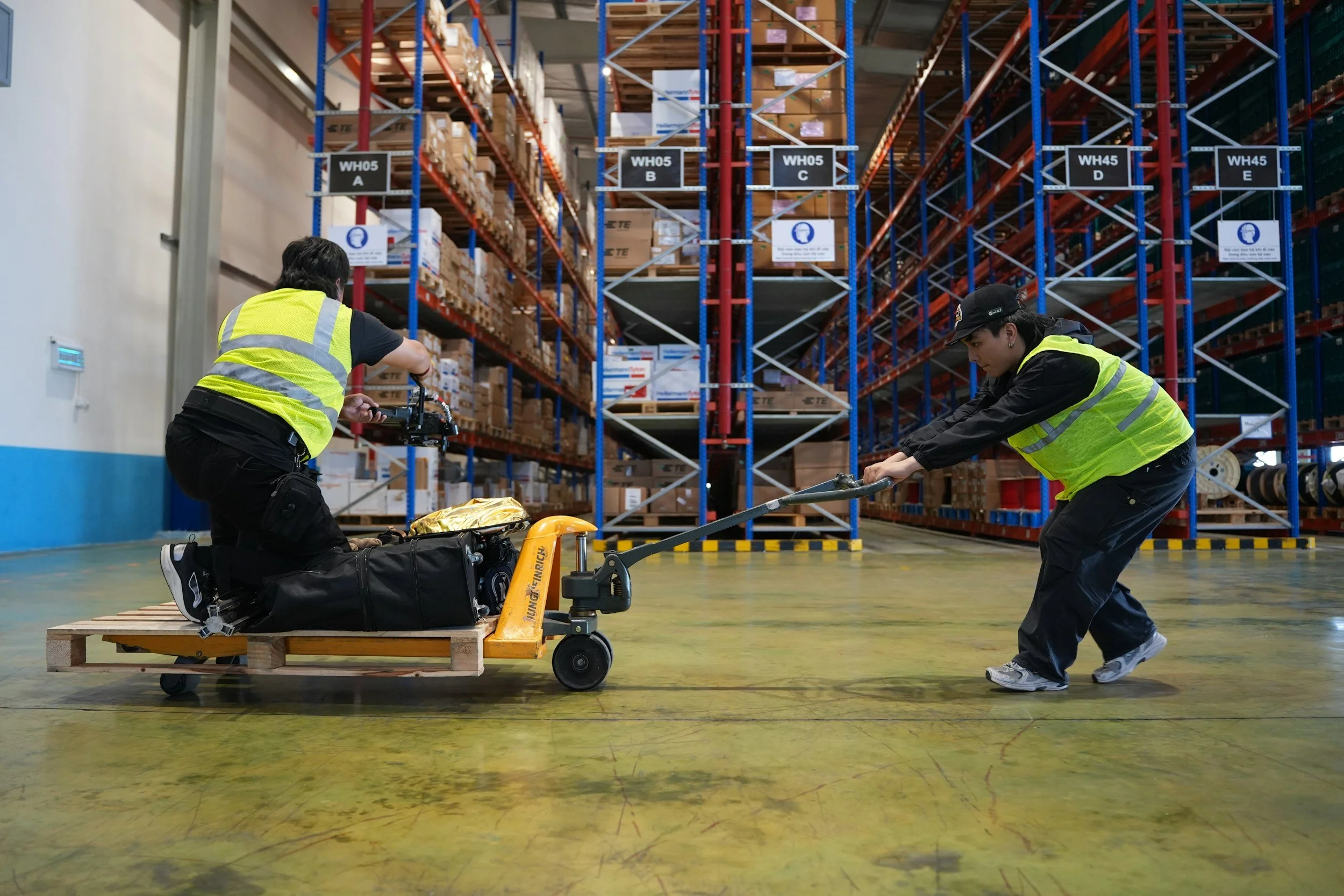 Two warehouse workers in safety vests pulling a pallet jack loaded with packages in a large storage facility with tall shelves filled with boxes.