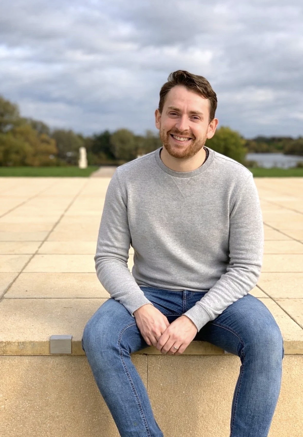 A smiling man sitting on a beige concrete step outdoors, with a cloudy sky and trees in the background.