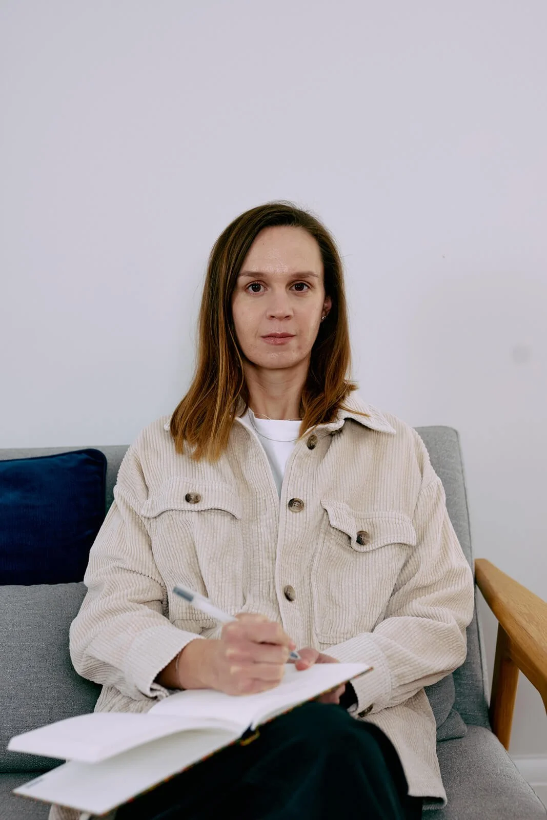 A psychotherapist with shoulder-length brown hair sitting on a grey sofa with a blue cushion, holding a pen and a notebook, looking at the camera in a room with white walls.