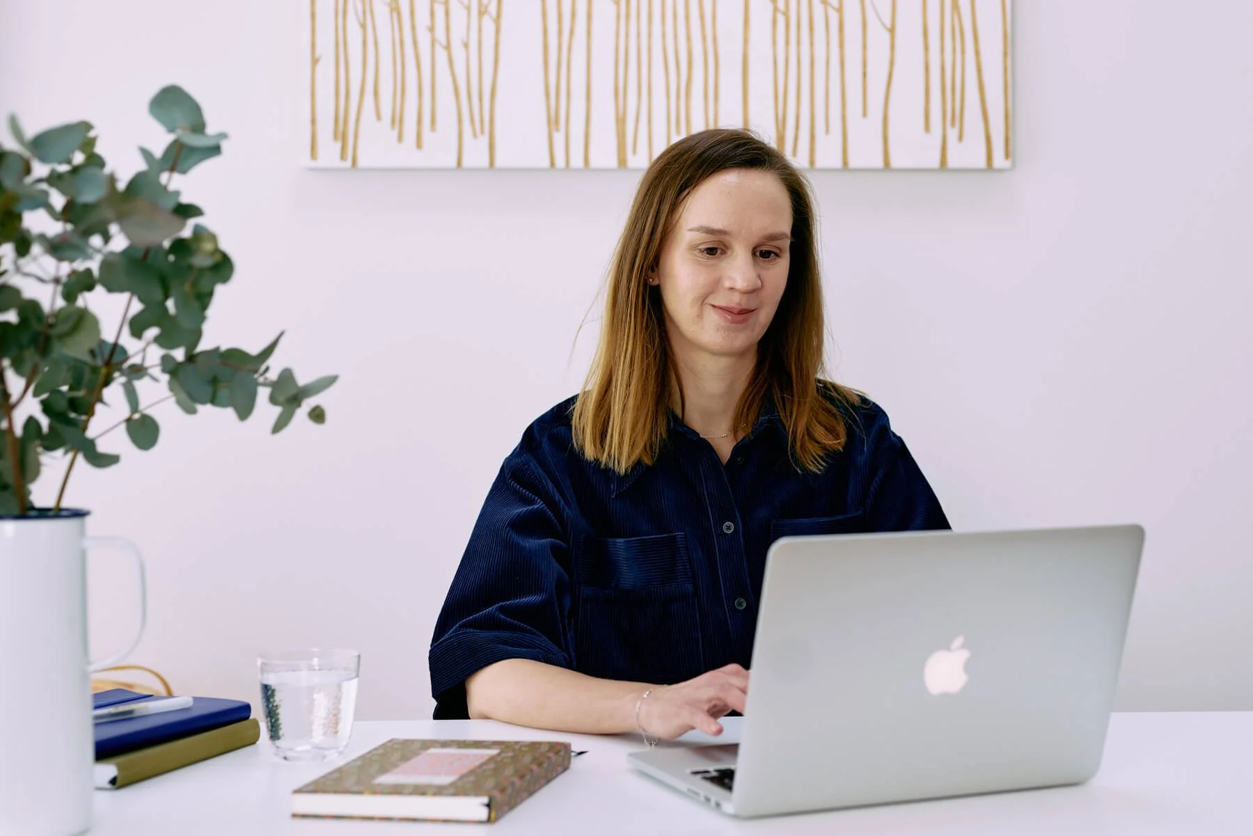 A counsellor with shoulder-length brown hair on a video call on a silver MacBook laptop at a white desk, with a plant, a glass of water, and notebooks on the desk, and a white wall with minimalist art in the background.